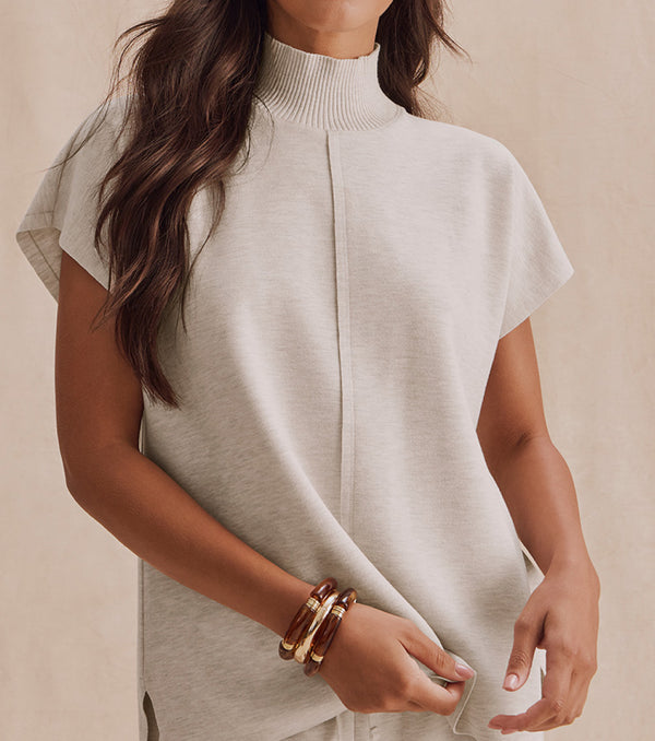 A woman wearing a light beige short-sleeve mock neck knit top, shown from the chest down against a neutral beige background.